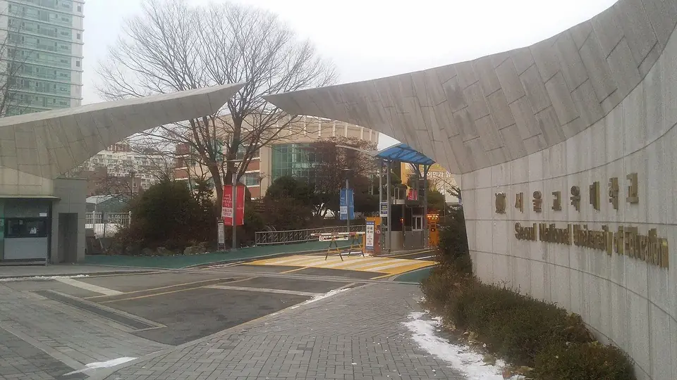 휘어진 지붕 모양의 문 구조물과 '서울교육대학교' 글자가 새겨진 벽, 그 뒤 나무와 건물