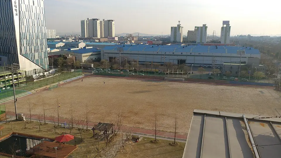 높은 곳에서 내려다본 흙 운동장과 그 너머 지붕이 파란 큰 건물들, 아파트가 보이는 풍경